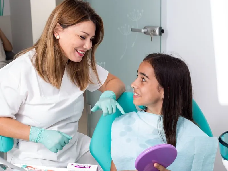 Medicul zâmbește unei fete care ține o oglindă în mână, în fotoliul dentar